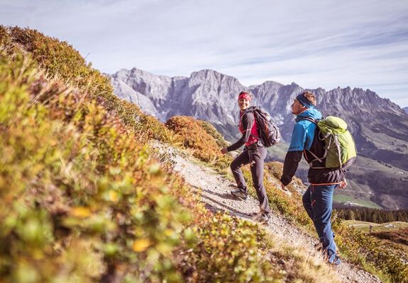 A woman and a man hike in the sunshine on a hiking trail with a view of the stony mountains of the Hochkönig massif in Salzburg