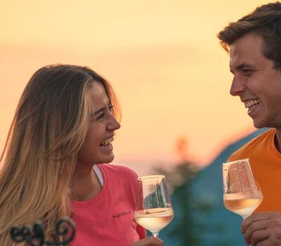 A man and a woman stand in the sunset in front of an alpine panorama, each with a glass of white wine in their hands