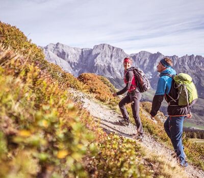 Eine Frau und ein Mann wandern im Sonnenschein auf einem Wanderpfad mit Ausblick auf das steinige Gebirge des Hochkönig Massivs in Salzburg