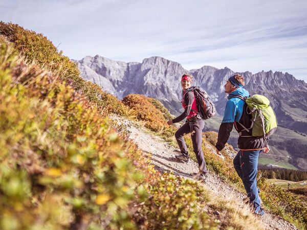 A woman and a man hike in the sunshine on a hiking trail with a view of the stony mountains of the Hochkönig massif in Salzburg