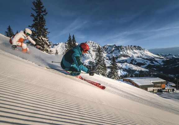 Zwei Skifahrer fahren auf einer frisch präparierten Skipiste im Sonnenschein im Skigebiet Mühlbach am Hochkönig