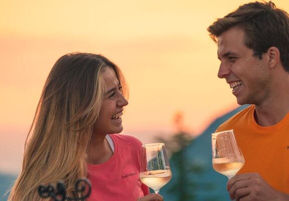 Ein Mann und eine Frau stehen im Abendrot vor einem Alpenpanorama mit jeweils einem Glas Weißwein in der Hand