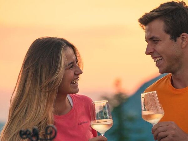 A man and a woman stand in the sunset in front of an alpine panorama, each with a glass of white wine in their hands